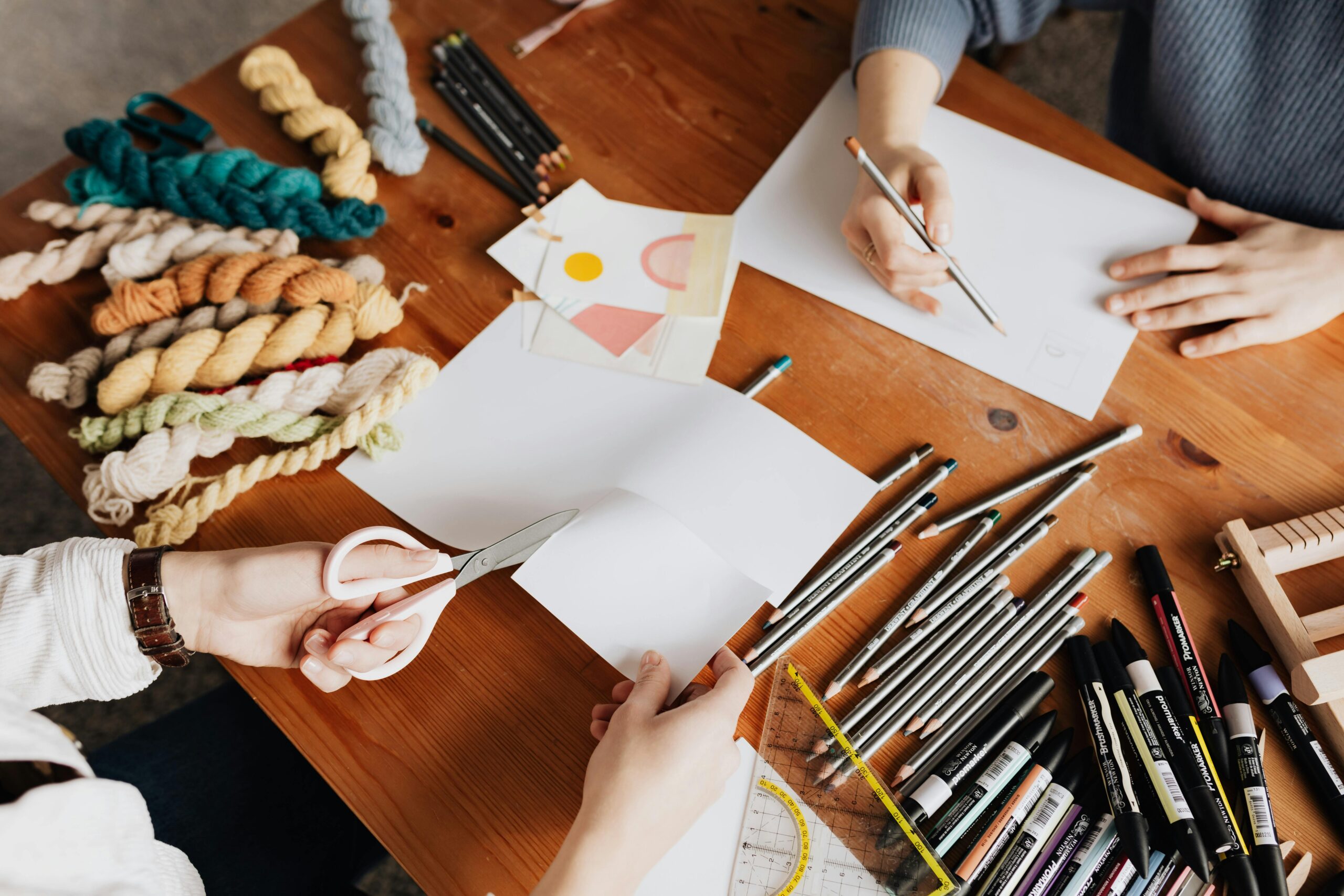 Artistic workspace with hands crafting and drawing using yarn, paper, and pencils.